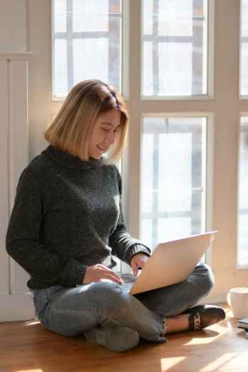 woman in gray sweater sitting on wooden floor typing on portable computer
